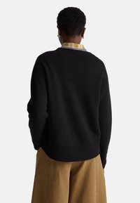 Schwarzer gestrickter Pullover mit einem gerippten Saum und Bündchen, getragen über einem gelben Karoshirt. Das Kleidungsstück hat eine lockere Passform und eine weiche Textur.
