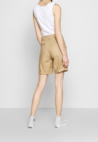 Béžové šortky s ohrnutými lemy, vyrobené z lehkého materiálu, v kombinaci s bílým topem bez rukávů a šedými vysoce špičkovými sportovními botami. Jednoduchý design, bez viditelných vzorů.