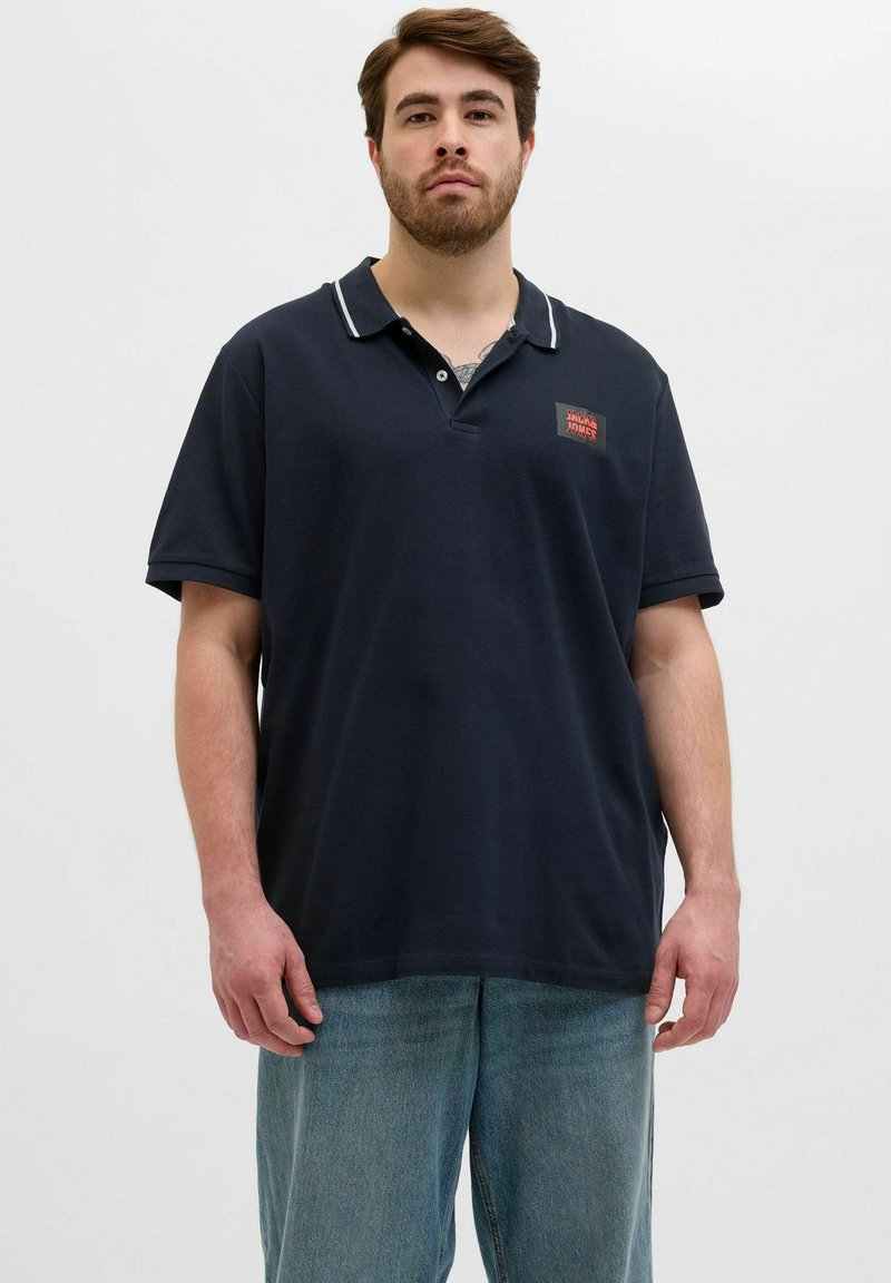 Navyblaues Polo-Shirt mit Kragen, weißen Akzenten und einem kleinen roten Logo auf der Brust. Kurze Ärmel und lockere Passform. Getragen mit hellblauen Jeans.