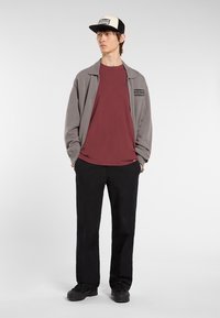 Mladý muž v šedé zipové bundě, vínovém tričku, černých kalhotách, černých botách a černo-bílém baseballovém klobouku, stojící s rukama v kapsách.