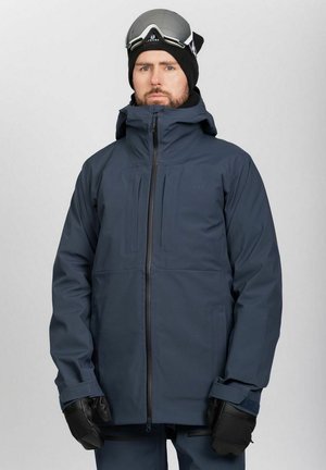 Mies, joka pukeutunut tummansiniseen talvitakkiin, mustiin hansikkaisiin, mustaan pipoihin ja jolla on laskettelulasit otsallaan, seisoo yksiväristä taustaa vasten.