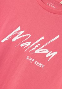Camiseta de algodón rosa con texto en blanco estilo pincel que dice "Malibu" y "SURF SHACK" debajo. Diseño clásico de cuello redondo.