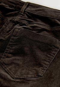 Schwarze Cordhosen mit einer Gesäßtasche, gestickten Details und einer weichen, strukturierten Oberfläche. Nahaufnahme betont den Stoff und die Verarbeitung.