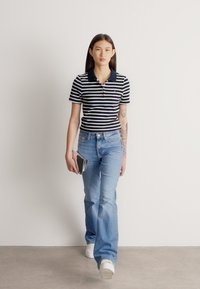 Csíkos galléros póló sötétkék és fehér színekben. Világoskék, bővülő szárú farmernadrág és fehér edzőcipő. A személy okostelefont tart a kezében.