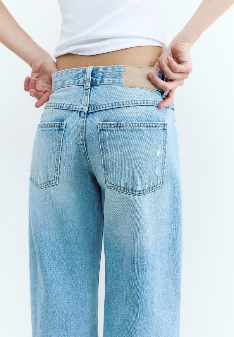 Háttal, mindkét kézzel a derékrészt igazítva egy világoskék magasderekú farmernadrágot és fehér felsőt viselő személy.