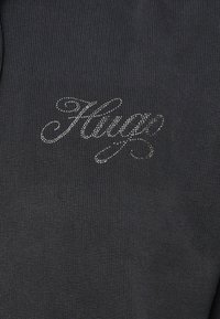 Sötét szövet bordás textúrával, ezüst hímzett logóval, amelyen a „Hugz” felirat látható egy ívelt betűtípussal.