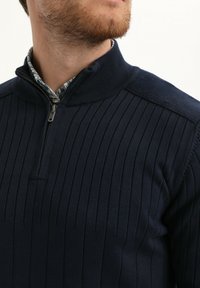 Navyblauwe gebreide pullover met een kwart-rits kraag, voorzien van verticale geribbelde textuur en contrasterende bloemenafwerking aan de binnenkant van de kraag.