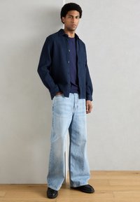 Sötétkék, gombos ing texturált mintával egy sötétkék póló fölött, világoskék, bőszárú farmernadrággal és fekete cipővel párosítva.