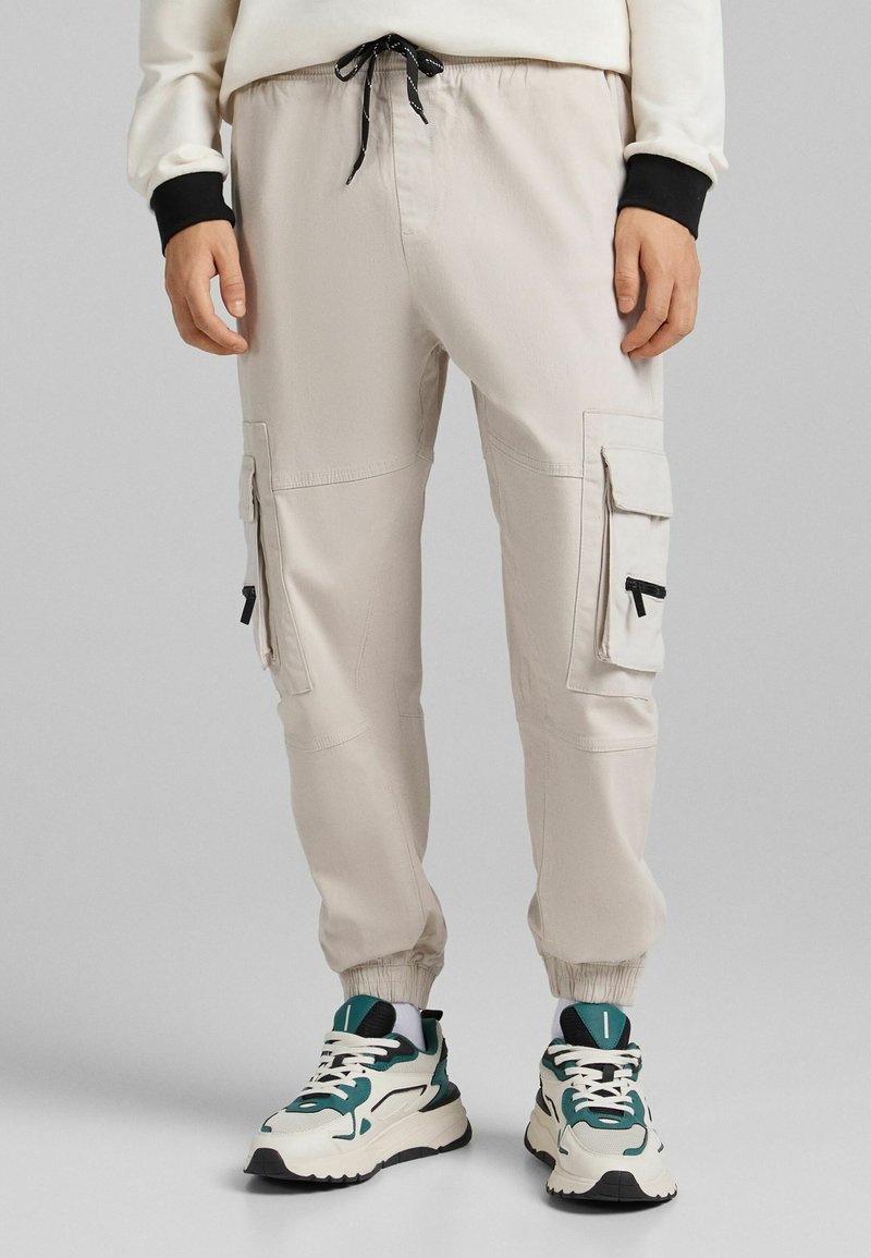 Osoba v béžových cargo kalhotách s elastickými manžetami a kapsami, doplněná bílými a tyrkysovými robustními teniskami.