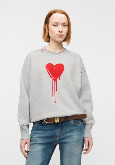 Γκρι φαρδιά φούτερ με κόκκινη καρδιά που στάζει. Συνοδεύεται από μπλε τζιν και καφέ ζώνη με χρυσή αγκράφα.