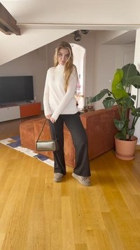 Μαύρο παντελόνι σε ευρεία γραμμή, λευκό πουλόβερ και γκρι παντόφλες. Κρατάει μια μικρή μαύρη τσάντα. Το φόντο περιλαμβάνει έναν καναπέ και μια γλαστρούλα.