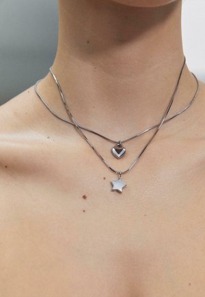 Dwa srebrne naszyjniki na szyi, jeden z zawieszką w kształcie serca nad drugim z zawieszką w kształcie gwiazdy na nagiej skórze.