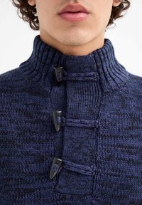 Suéter de punto azul marino con alto collar acanalado, que presenta tres botones de toggles negros y detalles trenzados en la parte frontal. Textura gruesa.