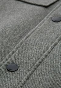 Sivi teksturirani materijal izbliza s gumbom s brendom "TOM TAILOR", istaknutim šavovima i krojenim dizajnom.
