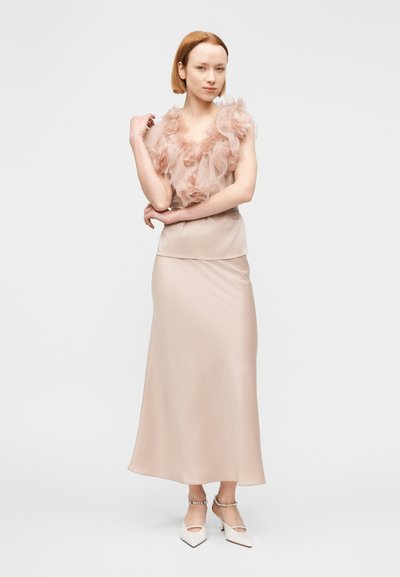 Robe longue et élégante en beige doux avec un haut en tulle texturé et superposé orné d'accents floraux. Escarpins blancs à bout carré et bijoux délicats.