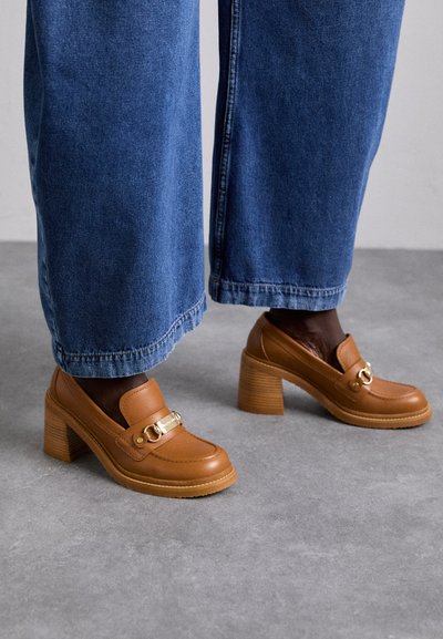 Mocasines de piel marrón con detalle de hebilla dorada, punta cuadrada y tacones de madera apilados, combinados con pantalones de mezclilla de pierna ancha en azul.