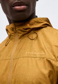 Primer plano de una persona que lleva una chaqueta Endura amarillo mostaza con cremallera y capucha, mostrando el logo en el pecho.