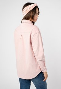 Roze fluwelen overhemd met een kraag, ruginzet en knoopmanchetten. Gecombineerd met een lichtroze fluwelen haarband. Achteraanzicht van de outfit.