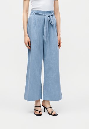 Femme portant un pantalon taille haute bleu clair à jambes larges avec une ceinture en tissu nouée, associé à des sandales à talons noires et un haut blanc.