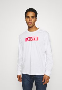 Camicia bianca a maniche lunghe con il logo rosso di Levi's sul petto; presenta un collo rotondo e un orlo dritto. Indossata con jeans in denim blu.