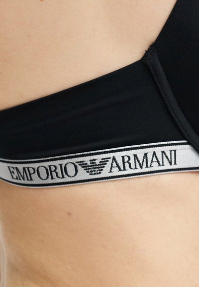 Posnetek od blizu strani pasu osebe, ki nosi črno spodnje perilo Emporio Armani z belo elastično obrobo z znamko.