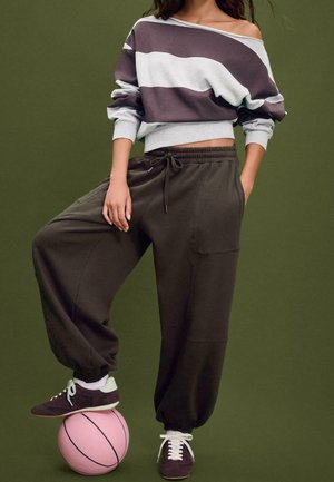 Personne en pantalon de jogging marron foncé ample et pull violet rayé épaules dénudées équilibre son pied sur un ballon de basket rose sur fond vert.