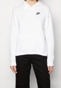 White hoodie with a front pocket, drawstring hood, and small black Nike logo. Soft fabric with ribbed cuffs and hem. Worn with black pants.