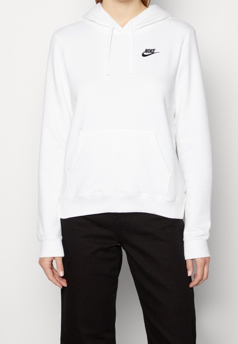 White hoodie with a front pocket, drawstring hood, and small black Nike logo. Soft fabric with ribbed cuffs and hem. Worn with black pants.