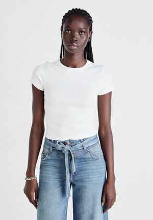 Kvinne med flettet hår iført en hvit tettsittende t-skjorte med korte ermer og høy midje blå jeans med knyttet belte, som står mot en enkel hvit bakgrunn.