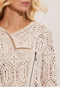 Virkningsjacka i krämfärg med geometriska mönster, sjalkrage och asymmetrisk dragkedjedetalj. Texturerad och öppenstickad design.