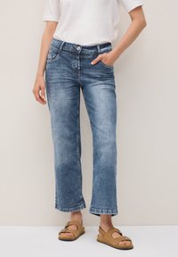 Une personne portant un t-shirt blanc et un jean bleu délavé se tient avec une main dans la poche, accompagnée de sandales marron sur un fond neutre.