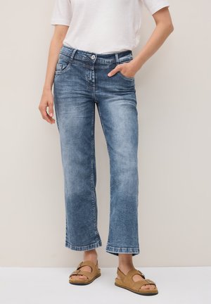 En person som bär en vit t-shirt och blekta blå jeans står med ena handen i fickan, tillsammans med bruna sandaler mot en neutral bakgrund.