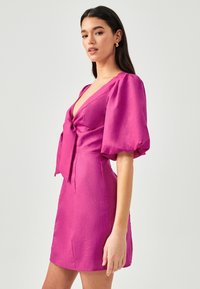 Fuchsia-mekko, jossa on syvä V-pääntie, sidottu nauhakoriste ja pussimaiset hihat. Valmistettu kevyestä kankaasta, jossa on sileä rakenne, ja se on yli polven pituinen.