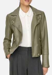 Giacca biker in ecopelle verde oliva con zip asimmetrica, colletto a scatto e dettagli trapuntati sulle spalle. Abbinata a una camicia bianca e pantaloni grigi.