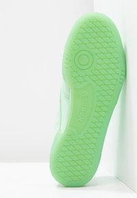 Grön gummisul med strukturerat mönster och cirkulär märkessymbol; har en mjuk övergång till en strukturerad kant.