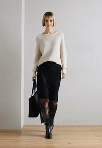 Off-white rebrasti pulover, crna čipkasta suknja s prozirnim rubom, crne gležnjače i crna torba za ramena čine slojeviti, teksturirani izgled.