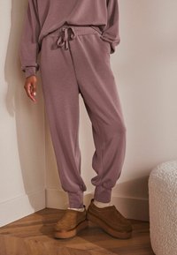 Vapaasti istuvat, mauve-väriset jogger-housut, joissa on elastinen vyötärö ja nauha, sivutaskut ja laskostetut nilkat, yhdistettynä ruskeisiin liukuverkkokenkiin.