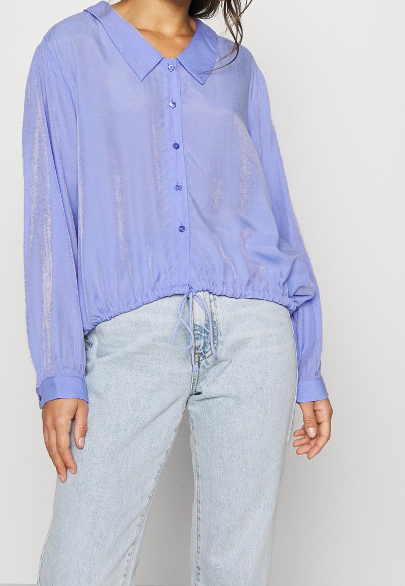 Femme portant un chemisier violet clair boutonné avec une taille à cordon et un jean bleu clair taille haute sur un fond uni.