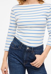 Γυναίκα με λευκή μπλούζα μακρυμάνικη με οριζόντιες μπλε ρίγες και σκούρο μπλε τζιν ψηλόμεσο, με το χέρι στην τσέπη.