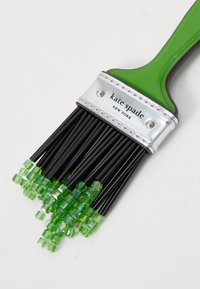 Brosse à cheveux noire avec un manche vert, une bande argentée métallique et des accents de perles vertes transparentes sur les poils, portant le nom de la marque "kate spade."