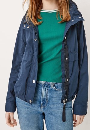 Osoba oblečená v tmavě modré bundě přes zelený žebrovaný svetr a světle modré džíny, stojící před neutrálním pozadím.