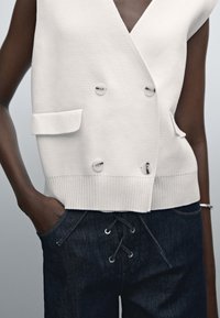 Biały dzianinowy bezrękawnik na guziki, noszony na ciemnych dżinsach z sznurowaniem, z jedną ręką w kieszeni i srebrną bransoletką na nadgarstku.