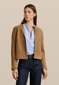 Kobieta w jasnobrązowej zamszowej kurtce z przeszyciami, pod którą ma niebieską bluzkę w pasiaki i ciemne dżinsy, spogląda w bok na jednolitym tle.