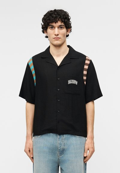 Άνδρας που φοράει μαύρο πουκάμισο με κουμπιά, με ριγέ μπλε και καφέ σχέδια στους ώμους και κείμενο "BARROW" στην τσέπη του στήθους.