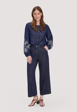 Frau steht, trägt eine dunkelblaue, geknöpfte Bluse mit weißer Blumenstickerei an den Ärmeln, weite Jeanshosen und braune Sandalen mit Absatz.