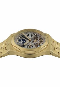 Goldfarbene Uhr mit einem Skelettzifferblatt, das komplexe Zahnräder zeigt. Sie verfügt über ein dickes, gewölbtes Gehäuse und ein robustes Metallarmband mit ineinandergreifenden Gliedern.