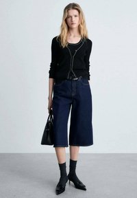 Črni dolg rokavnik z zadrgo, v kombinaciji z mornarskimi širokimi denim culottes. Model drži črno torbico; nosi temne nogavice in čevlje.