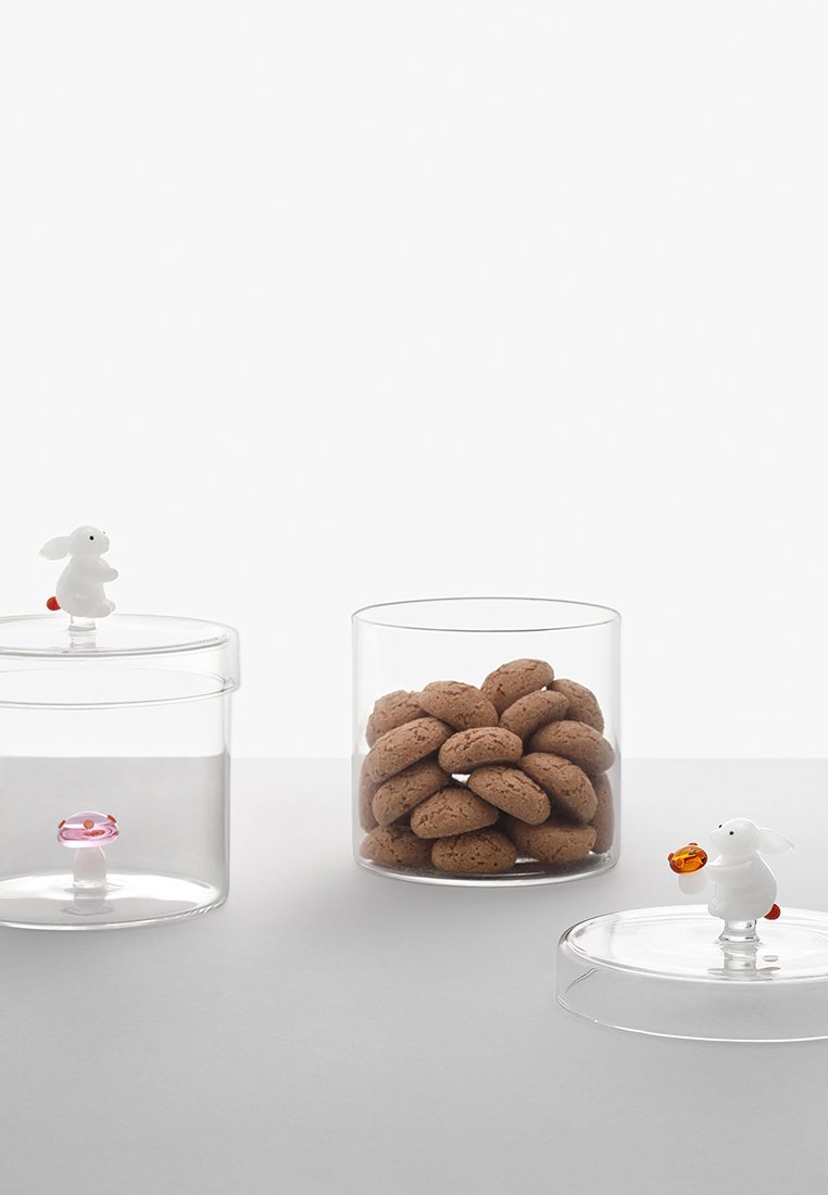 Dwa przezroczyste cylindryczne pojemniki z pokrywkami, jeden wypełniony brązowymi ciasteczkami, drugi pusty; pokrywki ozdobione małymi białymi figurkami królików trzymających różne przedmioty.