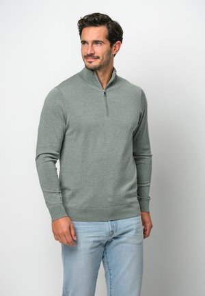 Hellgrüner Reißverschluss-Pullover aus weichem Stoff, mit hohem Kragen, langen Ärmeln sowie gerippten Bündchen und Saum, kombiniert mit hellblauen Jeans.