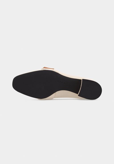 Semelle de chaussure habillée beige avec talon en caoutchouc noir et grip avant, affichant le nom de la marque embossé sur la zone de la voûte plantaire.